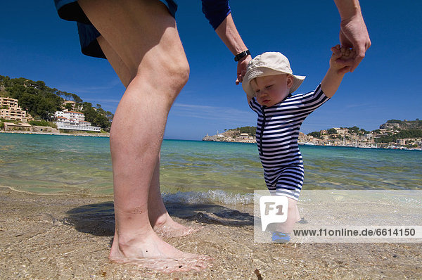 Father helping baby boy (6-11 months) walk on beach  Soller Majorca Balearic Islands Spain