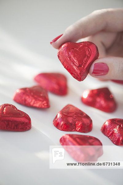 Close up of woman's fingers with red nail polish holding chocolate in shape of heart in red wrapping