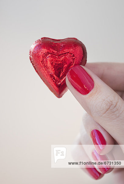 Close up of woman's fingers with red nail polish holding chocolate in shape of heart in red wrapping