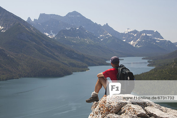 'Male Hiker With Backpack Sitting On A Rock Cliff Overlooking A Lake With Mountains And Blue Sky