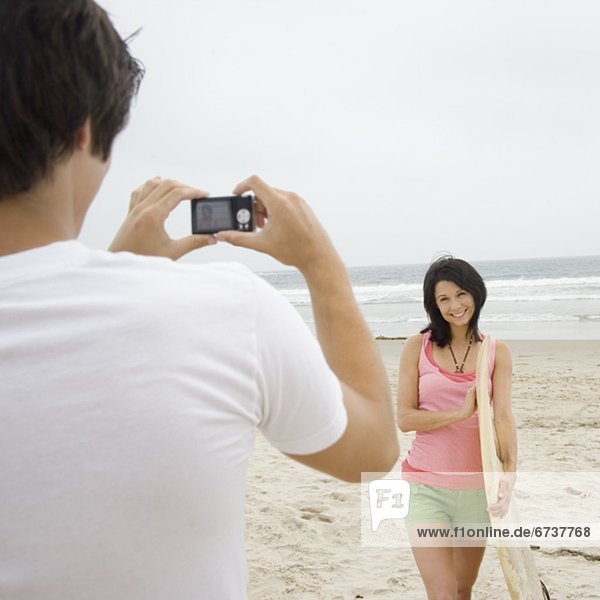 Man taking photograph of girlfriend with surfboard at beach