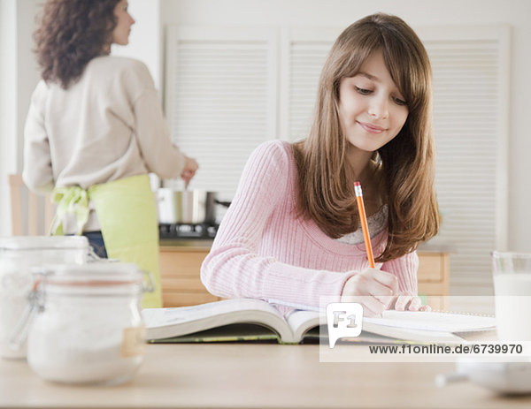Girl (10-12 years) doing homework in kitchen while her mother is cooking
