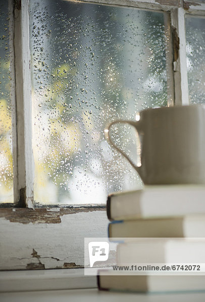 Cup on heap of books with rain on window in background