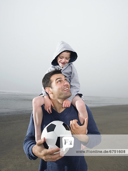Father carrying son on shoulders and holding soccer ball on beach