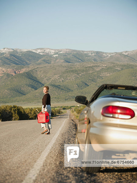 USA  Utah  Kanosh  Woman carrying canister walking along empty road  her car parked on roadside