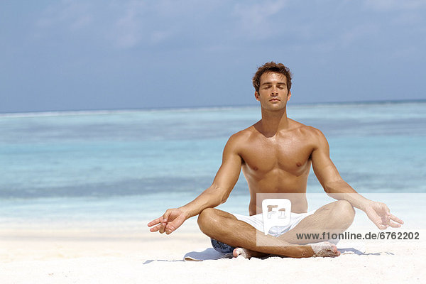 Handsome young man meditating in a lotus position on the beach