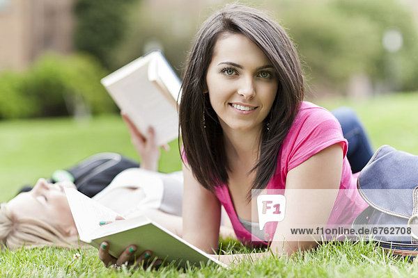Two female college student lying on grass reading books  one looking at camera