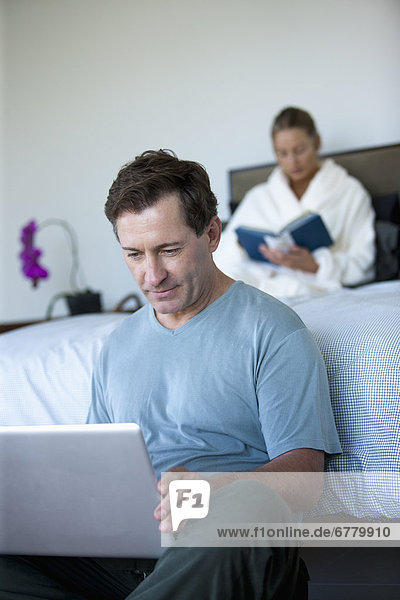 Mature man with laptop in bedroom and woman reading book in bed