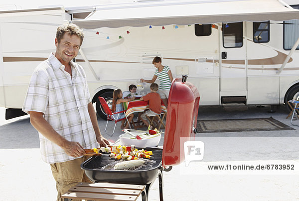 Man barbecuing with family and motor home in background