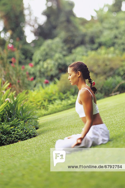 Frau sitzt auf Gras doing Yoga in einem tropischen Garten.