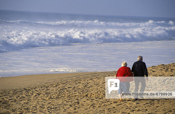 View from behind senior couple walking on beach  arm in arm white shorebreak ocean water
