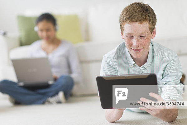 Boy (16-17) lying on floor using tablet pc  girl (14-15) with laptop in background