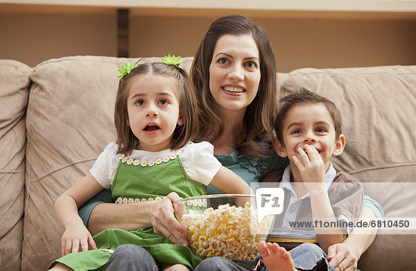 Front view of mother with children (4-5  6-7) sitting on couch watching tv and eating popcorn