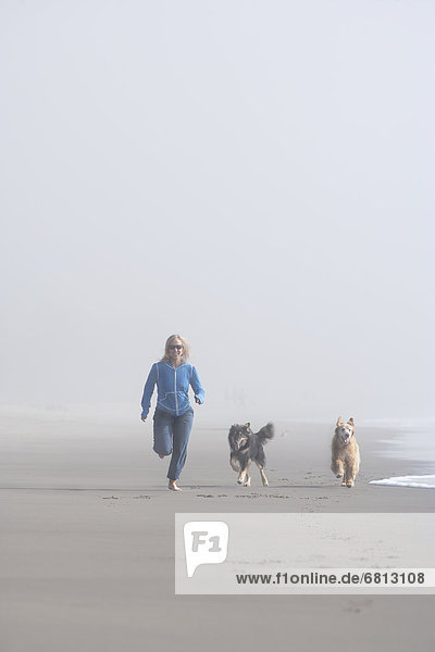 Woman running on sandy beach in fog with her dogs beside her