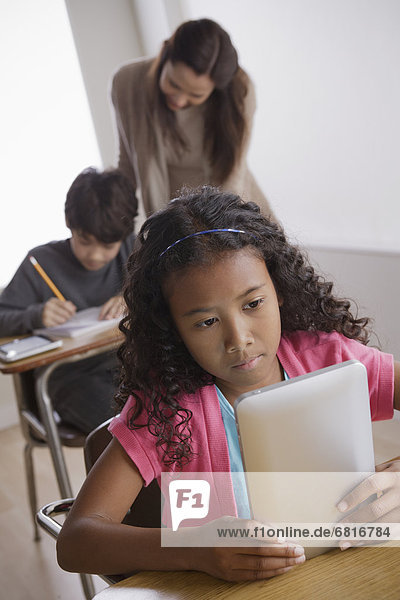Schoolgirl (10-11) holding digital tablet with boy (12-13) and teacher in background