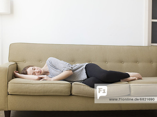 Junge Frau entspannt mit Buch neben sich auf braunem Sofa  lächelt