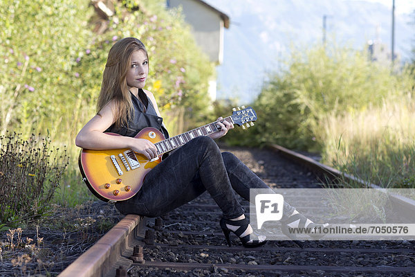 Junge Frau in Lederweste und Jeans posiert mit E-Gitarre auf Gleis
