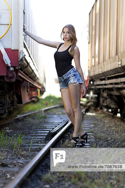 Junge Frau mit schwarzem Top  Jeans-Hotpants und hohen Schuhen posiert zwischen Güterwagons