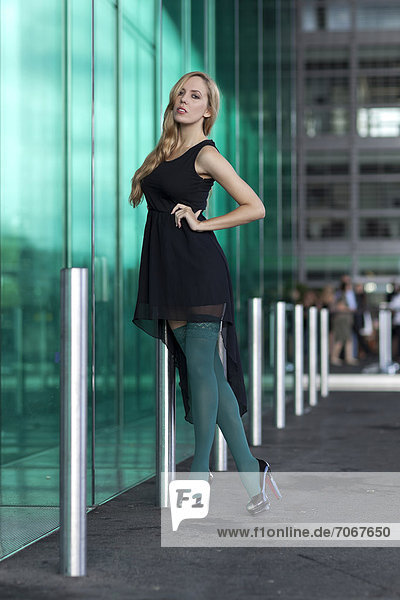 Young woman in a short black dress and green stockings posing in front of a green glass wall