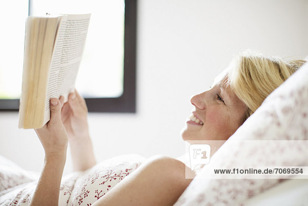Fröhliche mittlere erwachsene Frau auf dem Bett liegend und Buch lesend
