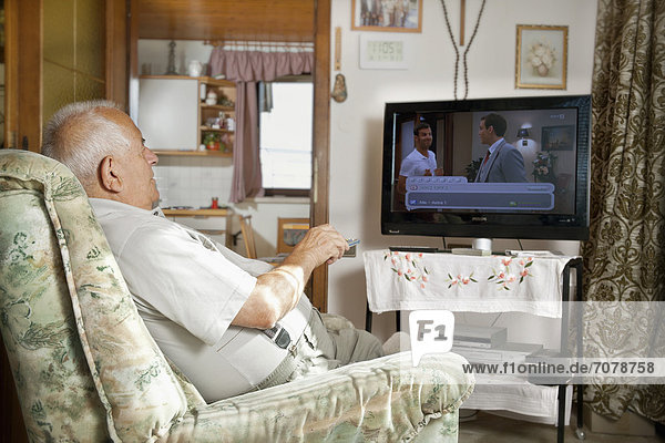 Elderly man sitting in front of a TV operating a remote control