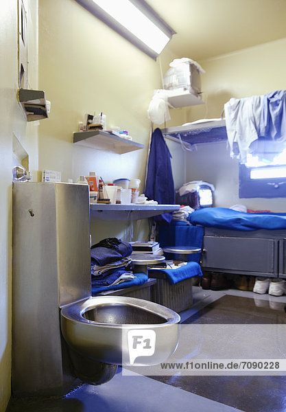 Prison cell. A small room  bed and urinal at a Correctional Facility. Fixed furniture and shelving. Inmate possessions. Jug and plate  personal belongings and clothes.