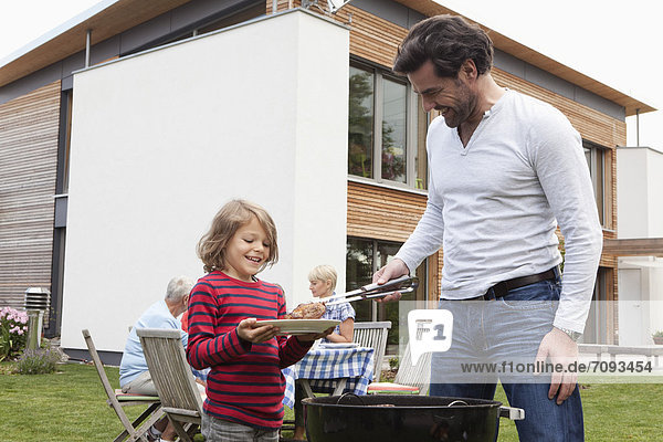 Germany  Bavaria  Nuremberg  Father and son preparing food on barbecue  family sitting in background