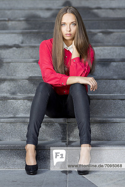 Young woman wearing a red top  black jeans and high heels posing while sitting on stairs