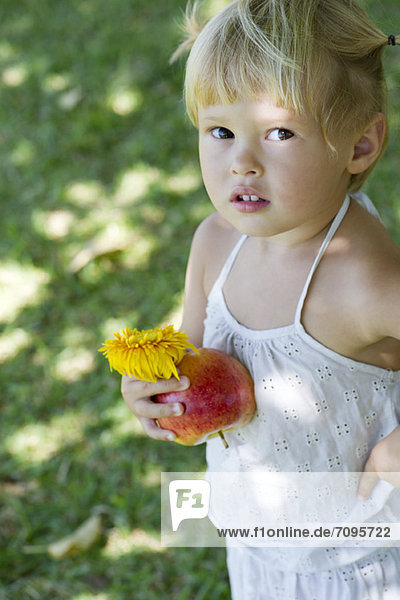 Kleines Mädchen  das draußen steht und Apfel und Blume hält