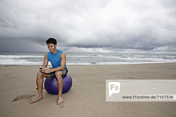 Young man sitting beach text messaging on cell phone
