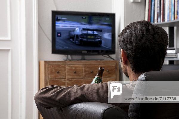 Man holding a bottle of beer while sitting on a leather sofa in front of a TV watching motorsport