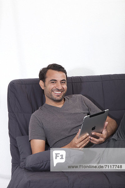 Smiling man lying on a gray sofa with an iPad