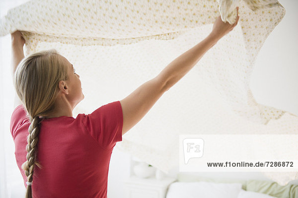 Young woman preparing clean sheet for bed