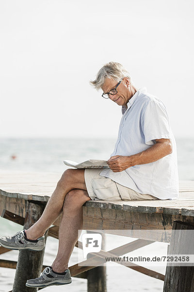 Spain  Senior man reading newspaper on jetty at the sea