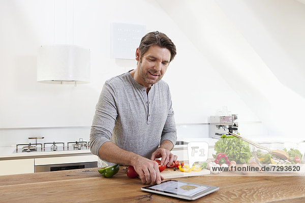 Mann,  der Essen zubereitet,  während er nach einer digitalen Tablette sucht