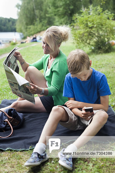 Reife Frau liest Zeitung sitzend mit Sohn mit Handy im Park