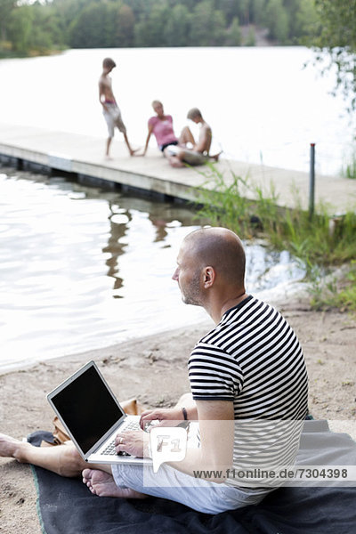 Der reife Mann schaut weg  während er den Laptop am Strand benutzt und die Familie am Pier sitzt.