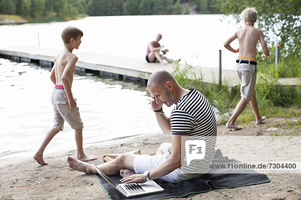 Reifer Mann mit Laptop bei der Kommunikation auf dem Handy am Strand mit der Familie im Hintergrund