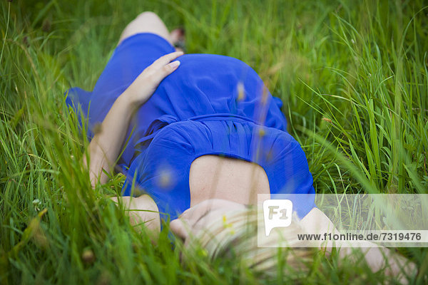 Junge schwangere Frau liegt im Gras und legt die Hand auf ihren Babybauch