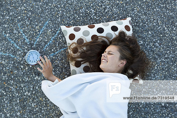 Junge Frau liegt mit Kissen und Decke am Boden  schlägt nach dem klingelnden Wecker  von oben aufgenommen