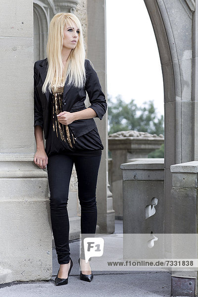 Junge Frau mit langen blonden Haaren  schwarzer Jacke und Hose posiert an Mauerbogen