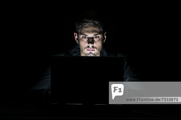 Junger Mann sitzt im Dunkeln vor seinem Laptop  nur vom Monitor angestrahlt
