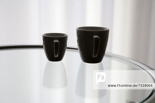 Eine Kaffeetasse und eine Espressotasse  die sich auf einem Glastisch spiegeln.