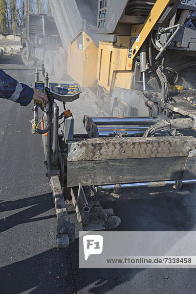 Manual worker controlling large road surfacing machine being trailed by articulated lorry
