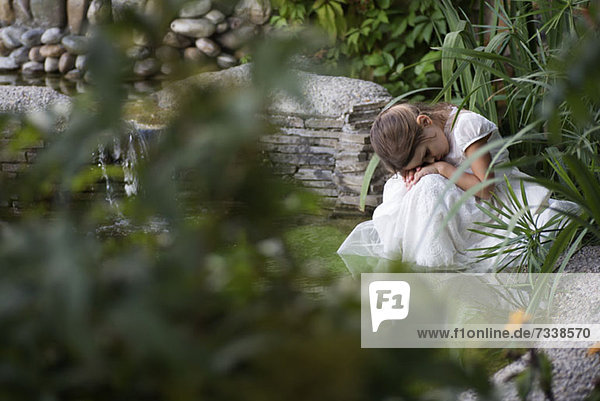 Ein junges trauriges Mädchen  das seinen Kopf auf die Hände legt  während es an einem Teich sitzt.