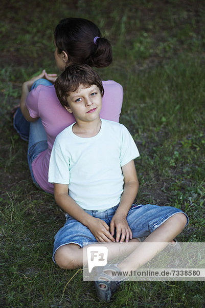 Ein Junge sitzt im Schneidersitz auf Gras und lehnt sich mit dem Rücken gegen den Rücken seiner Mutter.