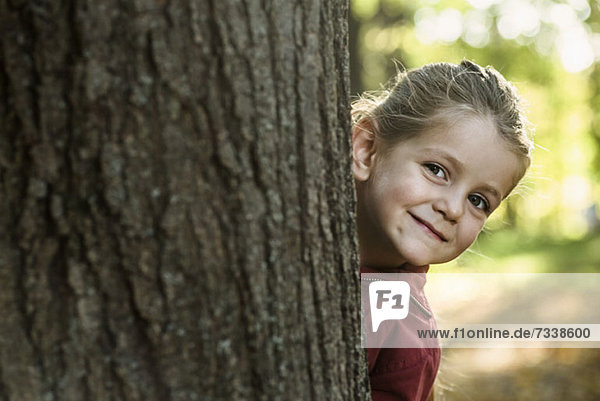 Ein junges lächelndes Mädchen  das hinter einem Baumstamm lugt.