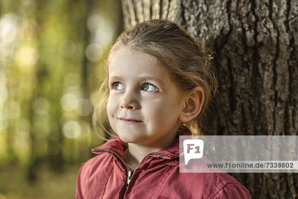 Ein junges Mädchen schaut neugierig weg  während es sich an einen Baumstamm lehnt.