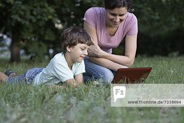 A mother and her son laughing while watching something on a laptop in a park
