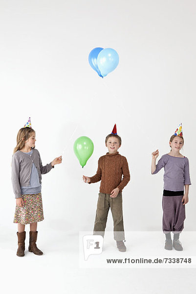 Drei Kinder tragen Partyhüte und halten Luftballons.